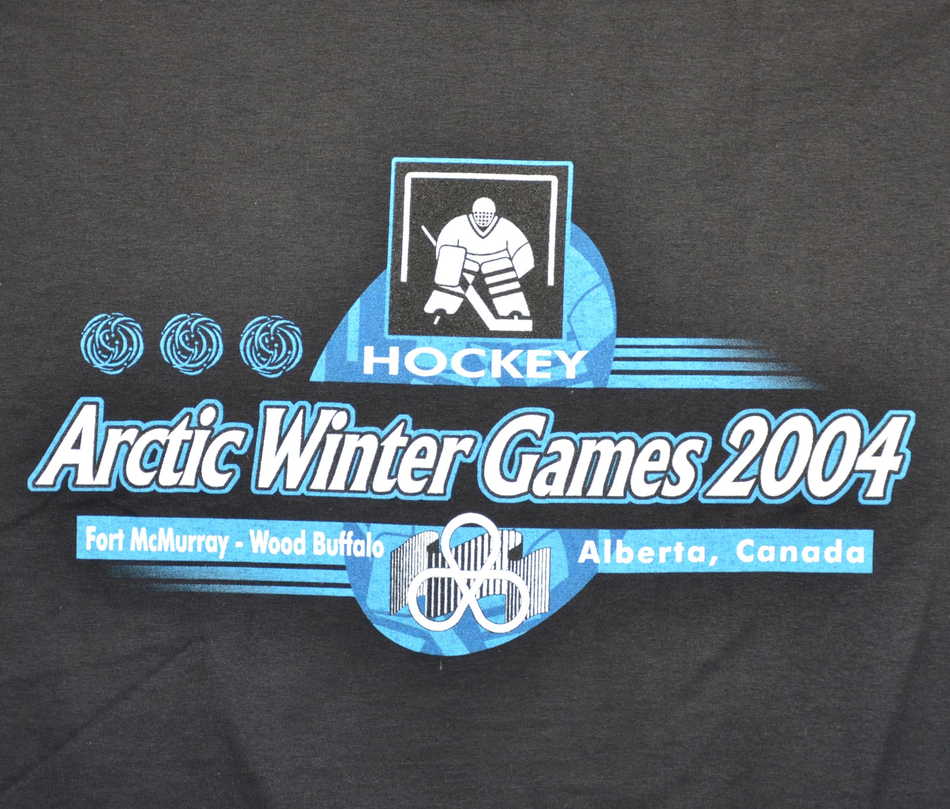 A black t-shirt featuring the Arctic Winter Games logo prominently displayed on the front.