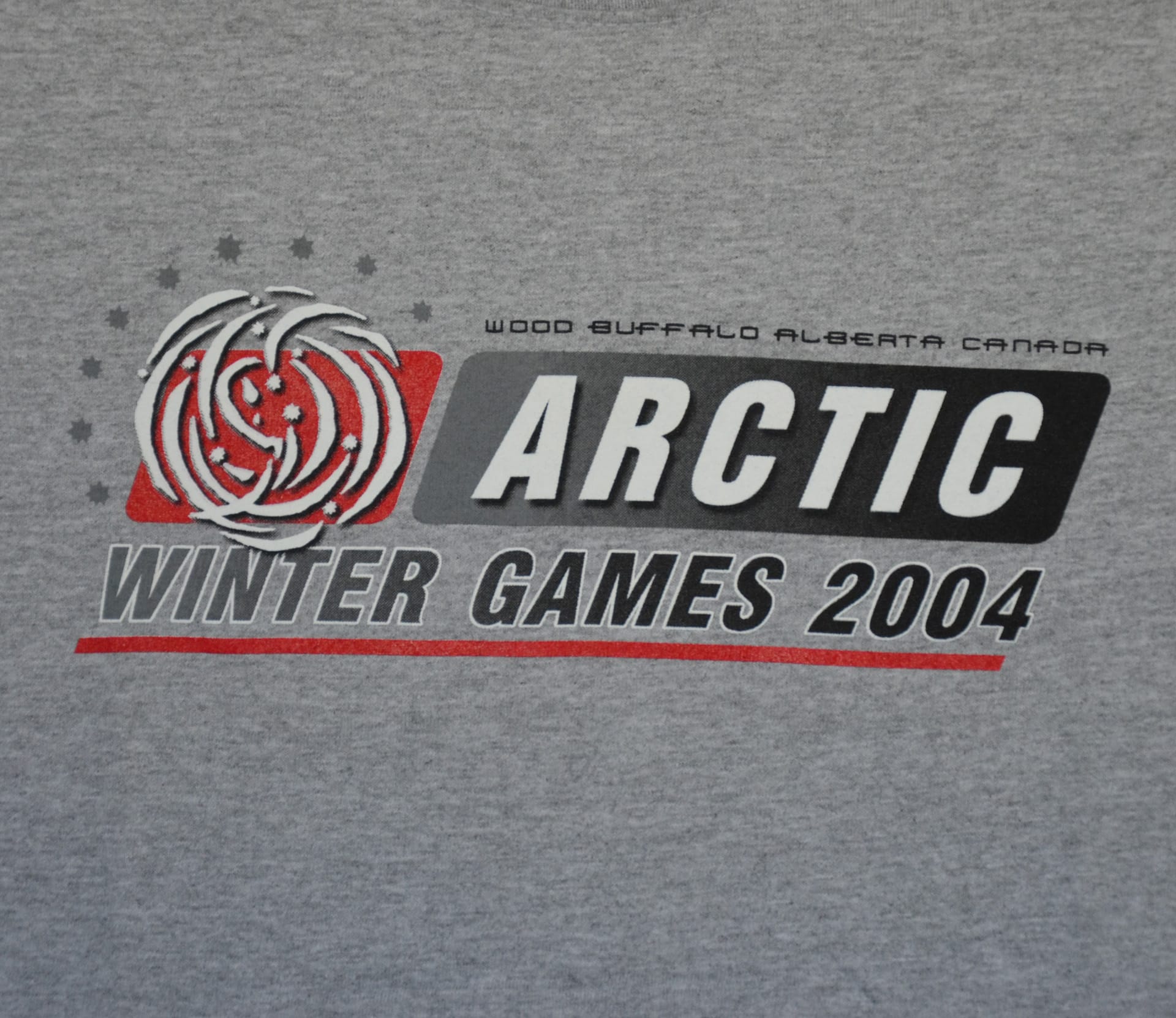 A grey t-shirt featuring the Arctic Winter Games logo prominently displayed on the front.